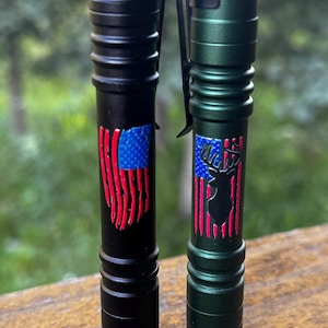 May include: Two flashlights, one black and one green, are displayed. The black flashlight features a distressed American flag design. The green flashlight has a deer silhouette over a similar flag design. Both have ribbed bodies.