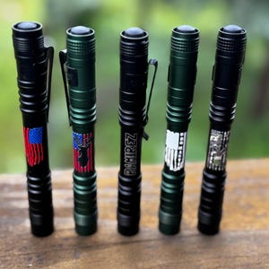 May include: Five black and green flashlights with pocket clips. The flashlights have various designs, including an American flag, a deer with an American flag, the name "RAMIREZ", a white flag design, and a circuit board design.
