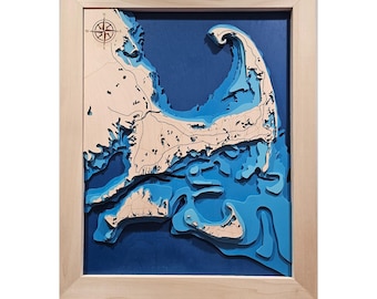 May include: A three-dimensional wooden bathymetric map of Cape Cod framed in light-colored wood. The map features layered cutouts representing land and varying depths of blue water, with a compass rose in the upper left corner and black lines.