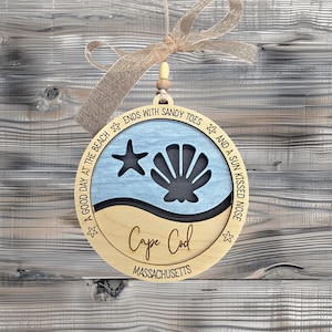May include: A wooden ornament with a blue and tan beach scene. The ornament features a seashell, a starfish, and the text "A good day at the beach ends with sandy toes and a sun kissed nose." The ornament also has the text "Cape Cod, Massachusetts" on it.