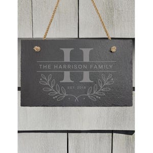 May include: A rectangular black slate sign with a rough, natural edge, suspended by a tan jute rope. The surface features an engraved large letter H, the text THE HARRISON FAMILY, a laurel leaf design, and the text EST. 2018 in a light gray color.