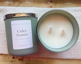May include: A sage green candle in a matte jar with a silver lid. The white label reads Whisper & Glow, Cider Season, apple, cinnamon, bourbon, coconut apricot wax, 12oz (340g). The candle has two wooden wicks and white wax.