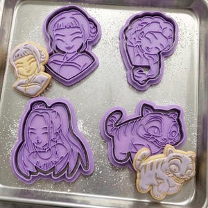 May include: A collection of purple plastic cookie cutters in various shapes, including a woman's face, a sleeping child, and a tiger. Two cookies are also present, matching the tiger and woman's face designs. The cookies are light brown with gold accents.