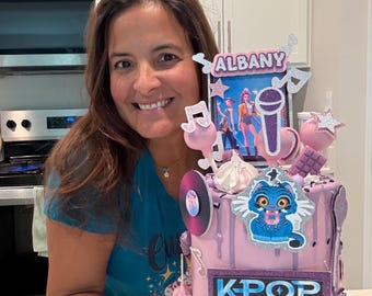 May include: A light purple tiered cake decorated with K-pop themed toppers including a sign that reads ALBANY, a microphone, musical notes, stars, a blue cat character, and a rectangular sign with the text KPOP DEMON HUNTERS in blue and white.