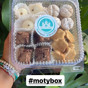 May include: A clear plastic container filled with a variety of small treats, including brownies, cookies, and white chocolate candies. The container has a sticker with the text "MOTYLAND" and a phone number. The text "#motybox #dulcesvenezolanos" is written below the container.