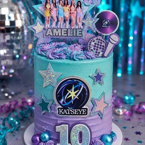 May include: A cylindrical cake with teal and purple ombre frosting, decorated with a Katseye logo, a number 10 topper, and a cutout of the group Katseye with the name Amelie. Additional accents include glittery stars, a disco ball, and a microphone.