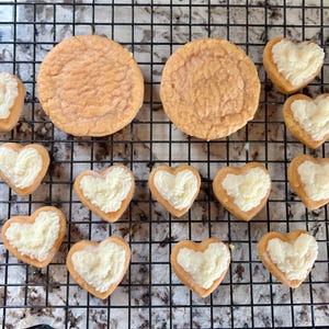 May include: Overhead shot of baked goods on a cooling rack. Two large round cookies and several heart-shaped cookies with white frosting are visible. The cookies are light brown in color.