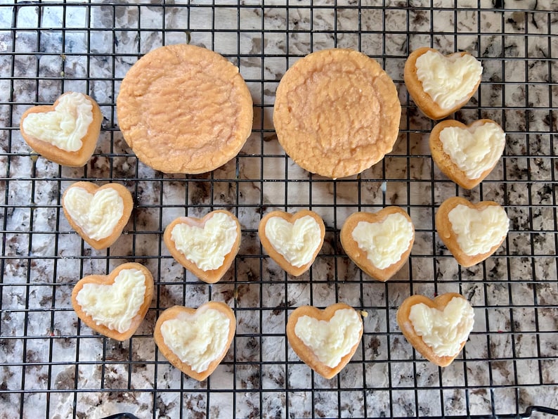 May include: Overhead shot of baked goods on a cooling rack. Two large round cookies and several heart-shaped cookies with white frosting are visible. The cookies are light brown in color.