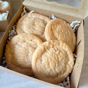 May include: A cardboard box with four round, light-brown baked goods. The baked goods have a textured surface and are nestled in white paper shreds. Another box with heart-shaped baked goods is visible in the background.