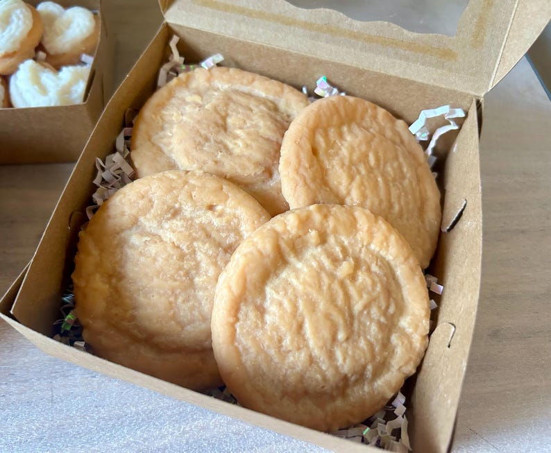 May include: A cardboard box with four round, light-brown baked goods. The baked goods have a textured surface and are nestled in white paper shreds. Another box with heart-shaped baked goods is visible in the background.