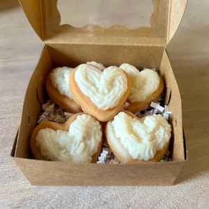 May include: A brown cardboard box with a clear window contains heart-shaped pastries. Each pastry has a light brown base and is topped with white frosting. The box is filled with white paper shreds.