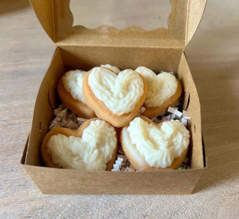 May include: A brown cardboard box with a clear window contains heart-shaped pastries. Each pastry has a light brown base and is topped with white frosting. The box is filled with white paper shreds.