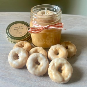 May include: A jar candle with a donut-shaped candle inside, tied with a red and white ribbon. The jar is next to a lid with text that reads "Craft Soy Candles & Woodwork Apple Cider Mini Donut With Love From MN". Several mini donut candles with white icing are in front.