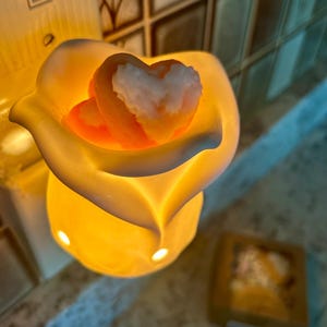 May include: A lit, yellow-orange wax warmer shaped like a flower, holding two heart-shaped wax melts. The melts are orange with white accents. The warmer is on a light-colored surface.