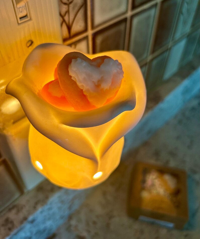 May include: A lit, yellow-orange wax warmer shaped like a flower, holding two heart-shaped wax melts. The melts are orange with white accents. The warmer is on a light-colored surface.