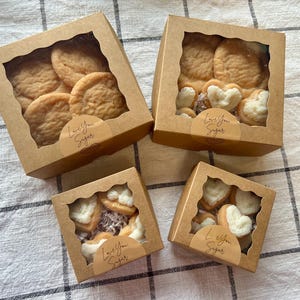 May include: Four kraft paper boxes with clear windows, each filled with cookies. Some boxes contain round cookies, others heart-shaped cookies. Each box has a sticker that reads "Love You Sugar". The cookies are arranged on a checkered surface.