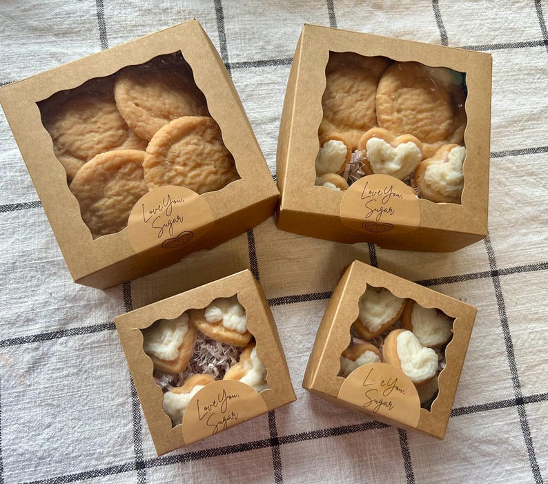 May include: Four kraft paper boxes with clear windows, each filled with cookies. Some boxes contain round cookies, others heart-shaped cookies. Each box has a sticker that reads "Love You Sugar". The cookies are arranged on a checkered surface.