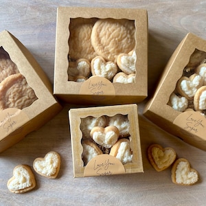 May include: Several boxes of cookies with clear window tops. Some boxes contain round cookies, others heart-shaped cookies with white filling. The boxes have a sticker that says "Love You Sugar".