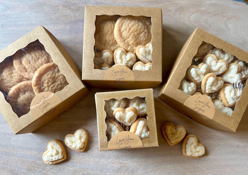 May include: Several boxes of cookies with clear window tops. Some boxes contain round cookies, others heart-shaped cookies with white filling. The boxes have a sticker that says "Love You Sugar".