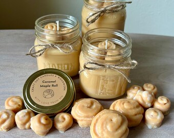 May include: Three clear glass mason jars filled with light brown soy wax candles, each tied with a brown twine bow. A gold metal lid sits nearby, labeled "Craft Soy Candles & Woodwork, Caramel Maple Roll, With Love From MN." Small wax melts shaped like cinnamon rolls are scattered around the jars.