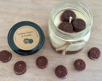 May include: A soy candle in a glass jar topped with three dark brown cookie-shaped wax embeds, tied with twine. The black lid features a tan label reading Craft Soy Candles & Woodwork, Cookies, Cream, & Crumbs, With Love From MN. Six small cookie-shaped wax pieces surround the jar.