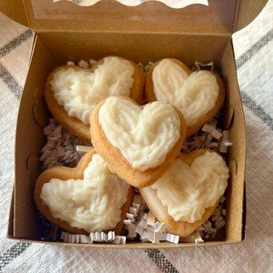 May include: A cardboard box filled with heart-shaped cookies. The cookies have a golden-brown base and are topped with white frosting. The box is lined with white and silver crinkle paper.