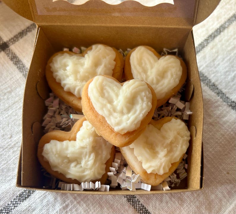 May include: A cardboard box filled with heart-shaped cookies. The cookies have a golden-brown base and are topped with white frosting. The box is lined with white and silver crinkle paper.