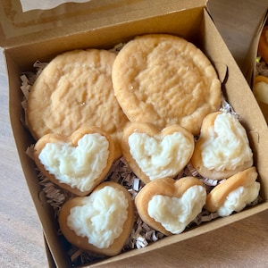 May include: A box of baked goods, including two large round cookies and several heart-shaped cookies with white frosting. The cookies are arranged in a brown cardboard box with crinkle paper.