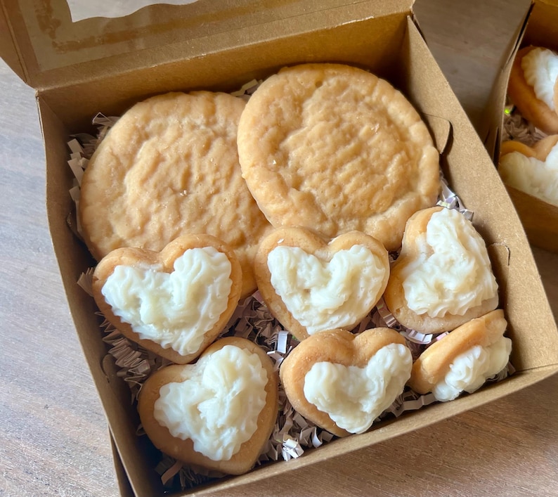 May include: A box of baked goods, including two large round cookies and several heart-shaped cookies with white frosting. The cookies are arranged in a brown cardboard box with crinkle paper.