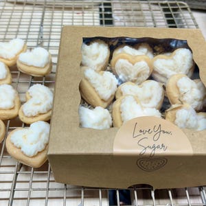 May include: Heart-shaped cookies with white frosting, some arranged on a metal cooling rack and others in a cardboard box with a clear window. The box has a sticker that says "Love You, Sugar."