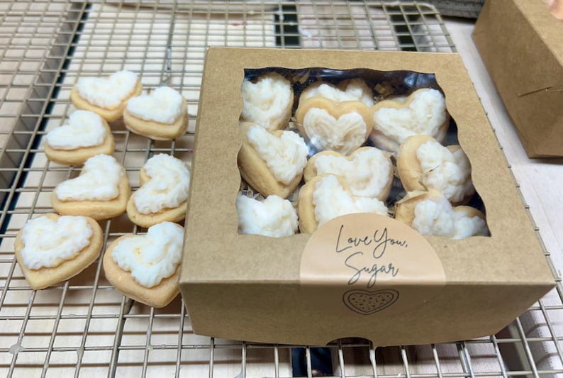 May include: Heart-shaped cookies with white frosting, some arranged on a metal cooling rack and others in a cardboard box with a clear window. The box has a sticker that says "Love You, Sugar."