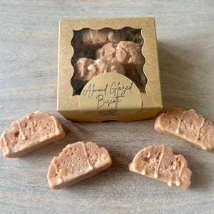 May include: A box of almond glazed biscotti with a clear window, alongside several individual biscotti. The biscotti are light brown with a white glaze drizzle. The box has a label that reads "Almond Glazed Biscotti."