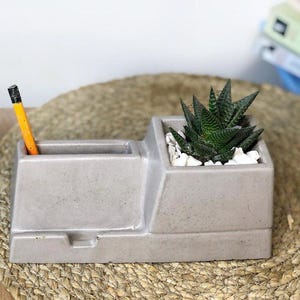 May include: A grey concrete desk tidy with a built-in planter and pencil holder. The planter contains a green succulent and white pebbles. A yellow pencil with a black eraser is in the pencil holder.