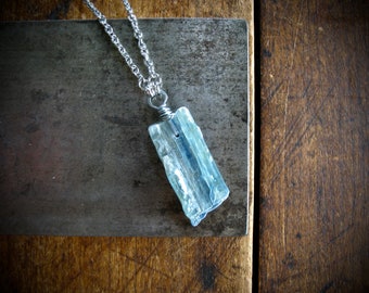 May include: A rectangular, light blue crystal pendant necklace with a silver chain. The crystal is wrapped in silver wire and hangs against a textured, gray metal surface and a wooden background.