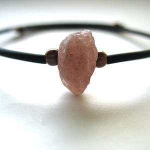 May include: A black bracelet with a rough, pink gemstone centerpiece. The gemstone is held in place by two small, bronze-colored beads. The bracelet is a simple, elegant design.