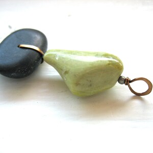 Op de afbeelding: Handgemaakte hanger met een gladde, donkergrijze steen verbonden met een lichtgroene steen met een bronskleurige draad. De hanger heeft een bronzen lus om op te hangen. De stenen liggen op een wit oppervlak.