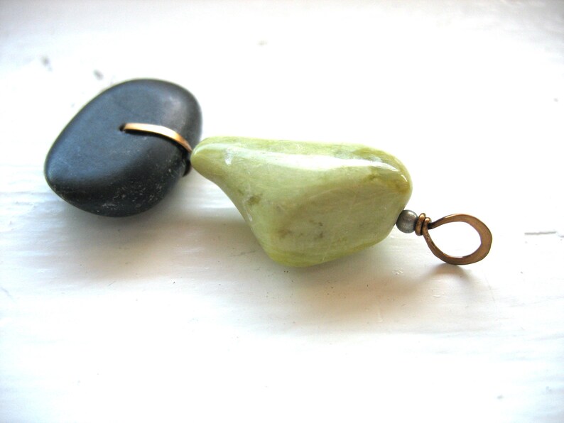 Op de afbeelding: Handgemaakte hanger met een gladde, donkergrijze steen verbonden met een lichtgroene steen met een bronskleurige draad. De hanger heeft een bronzen lus om op te hangen. De stenen liggen op een wit oppervlak.