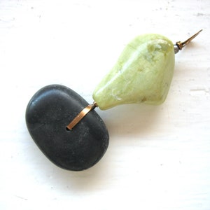 Op de afbeelding: Een unieke, handgemaakte hanger met een zwarte steen en een lichtgroene steen, verbonden door koperkleurig metaal. De groene steen heeft een puntig metalen element. De hanger staat tegen een witte achtergrond.