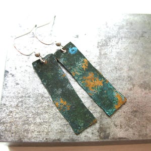 May include: A pair of handmade earrings with a rectangular shape. The earrings have a distressed, textured appearance with a green and copper color scheme. The earrings hang from silver-colored ear wires.