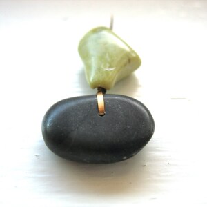 Op de afbeelding: Een handgemaakte ketting met een gladde, donkergrijze stenen hanger en een lichtgroene steen. De stenen zijn verbonden door een goudkleurige metalen ring, wat een uniek, minimalistisch ontwerp cre&euml;ert.