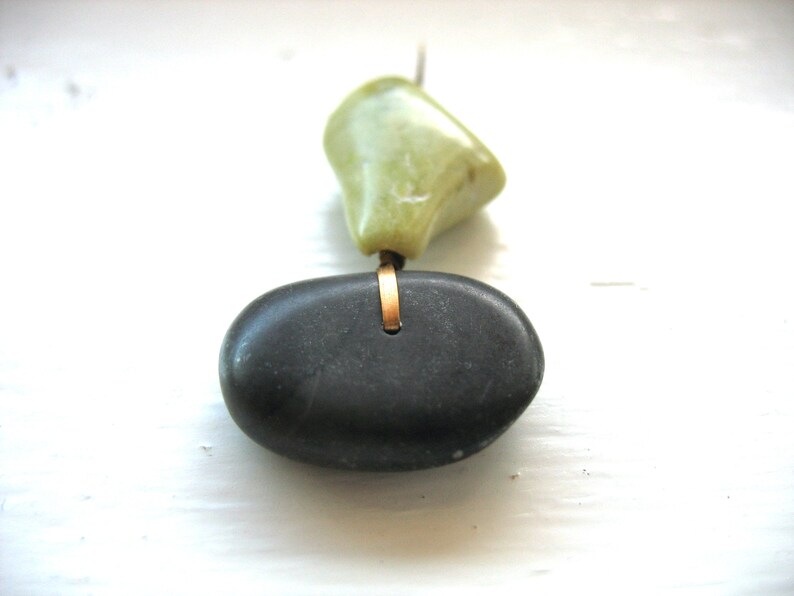 Op de afbeelding: Een handgemaakte ketting met een gladde, donkergrijze stenen hanger en een lichtgroene steen. De stenen zijn verbonden door een goudkleurige metalen ring, wat een uniek, minimalistisch ontwerp cre&euml;ert.