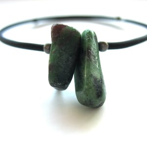 May include: A necklace featuring a thin black cord and two tumbled green and dark red stones. The stones are positioned side by side at the center, held in place by small metallic beads on either side of the stones along the black cord.
