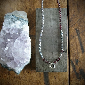 Op de afbeelding: Een ketting met een streng kleine grijze parels en donkerrode granaatkralen, in het midden voorzien van een enkele heldere druppelvormige hanger. Naast de ketting ligt een groot, grillig stuk lichtpaars amethistkristal.