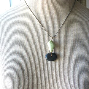 Op de afbeelding: Zilveren ketting met een hanger. De hanger heeft een lichtgroene, onregelmatig gevormde steen boven een zwarte, ovaalvormige steen. Een dunne, goudkleurige staaf verbindt de twee stenen. De ketting wordt getoond op een beige mannequin.