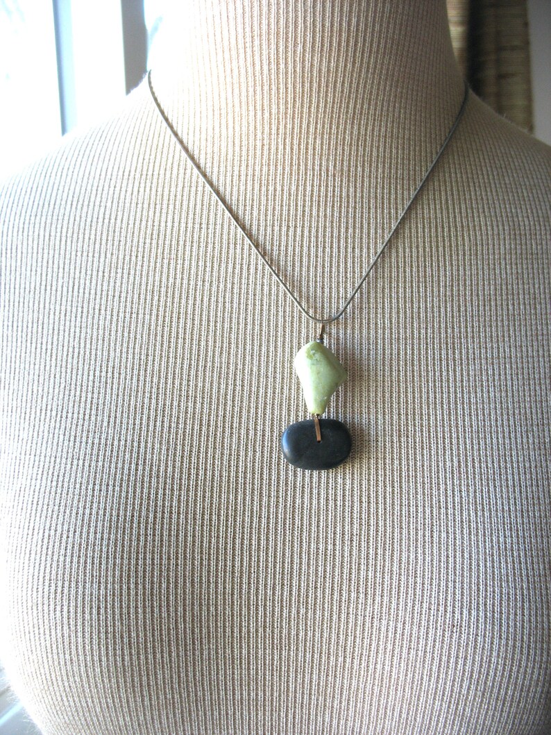 Op de afbeelding: Zilveren ketting met een hanger. De hanger heeft een lichtgroene, onregelmatig gevormde steen boven een zwarte, ovaalvormige steen. Een dunne, goudkleurige staaf verbindt de twee stenen. De ketting wordt getoond op een beige mannequin.
