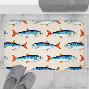 May include: A rectangular bath mat featuring a repeating pattern of cartoon fish. The fish have blue bodies, orange tails, and yellow fins, set against a cream background. Gray slippers are visible in the lower left corner.