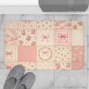 May include: A rectangular bath mat with a patchwork design in shades of pink and cream. The mat features floral patterns, bows, and a gingham print. Gray slippers are at the bottom of the mat.