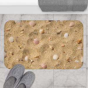 May include: Rectangular bath mat featuring a beach scene with sand and seashells. The mat has a light brown background with various seashells in shades of white, pink, and tan. Gray slippers are at the bottom.