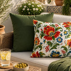 May include: A square throw pillow featuring a white background with a vibrant floral pattern in red, orange, blue, and green, placed in front of a solid dark olive green square throw pillow on a light-colored outdoor sofa cushion.