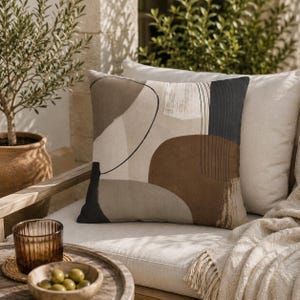 May include: Decorative throw pillow with an abstract design in shades of brown, beige, and black. The pillow features a geometric pattern with curved lines and shapes. It is placed on a white cushion with a beige blanket.